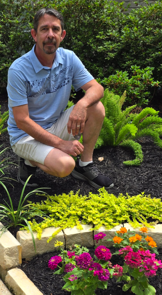 Neil James kneeling in landscaped garden bed with colorful flowers
